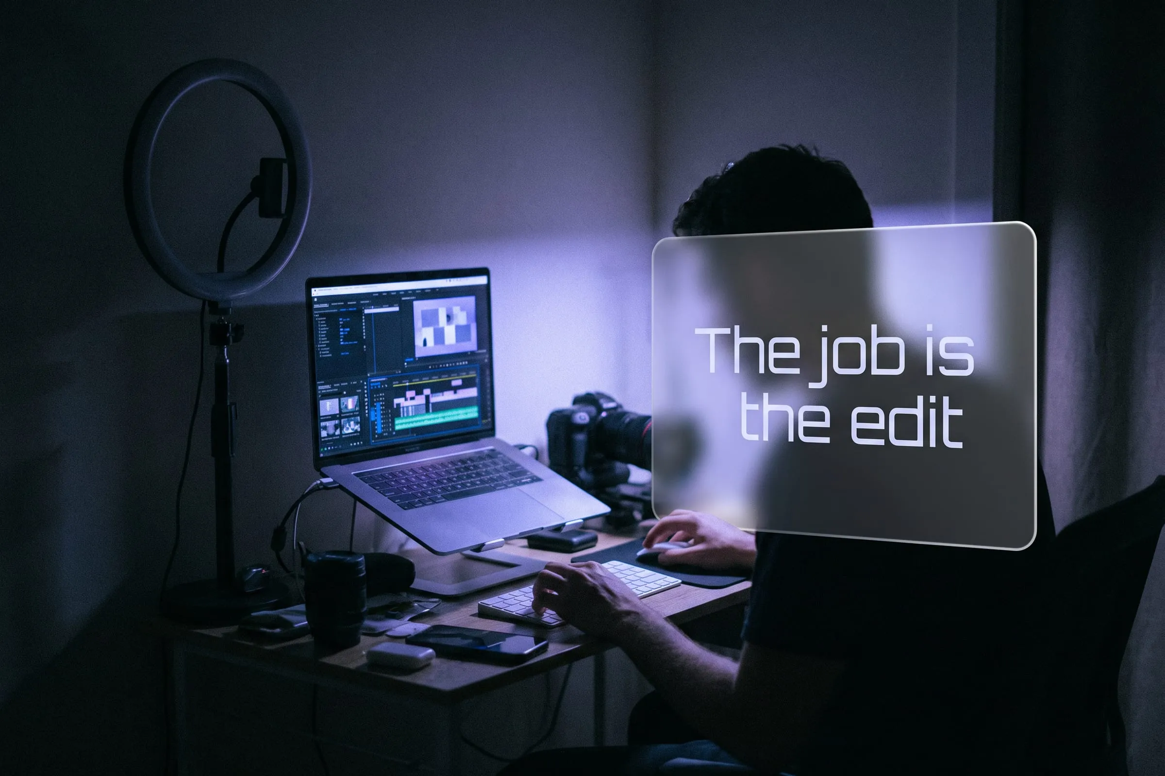 A person edits a video timeline on a laptop at a home desk, with an unlit ring light and camera behind them.