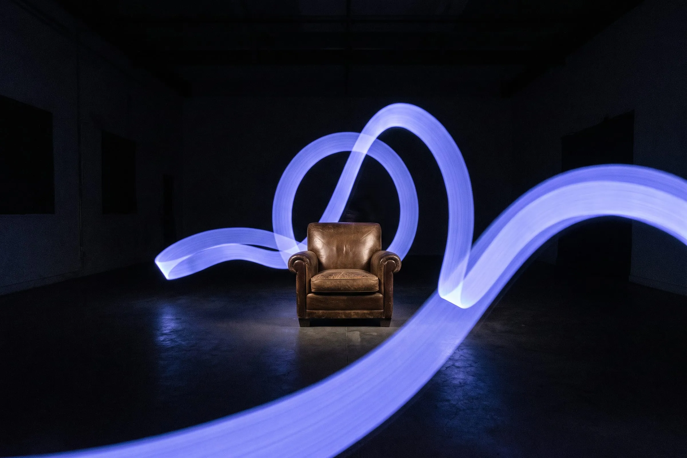 An empty armchair alone in a bare room with light trails sweeping around it.