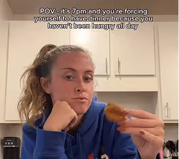 Woman in a blue hoodie sitting in a kitchen at 7pm, reluctantly holding food with text overlay about forcing oneself to eat dinner when not hungry.