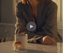 A person wearing a dark sweater sitting at a table with a cup and a small hamster or mouse nearby.