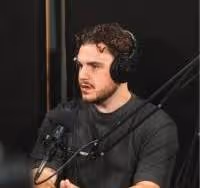 Man with curly hair wearing headphones and speaking into a microphone in a recording studio.